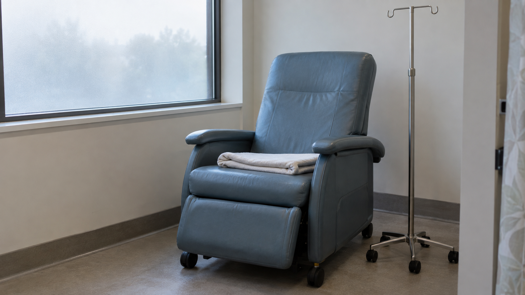 An empty blue treatment recliner with a folded blanket beside an IV pole and frosted window.